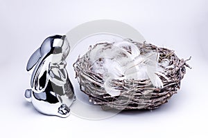 Small and cute silver easter bunny figure looking into a empty easter nest made of branches, filled up with white feathers, white
