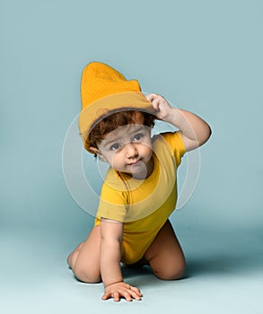 Small cute smiling curly baby boy toddler in yellow comfortable jumpsuit and hat crawling on floor