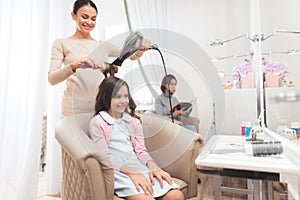 A small dark-haired girl sits in the beauty salon chair. The hairdresser dries the hair to the girl with a hairdryer.