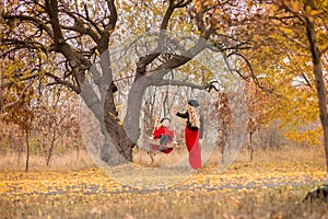 Beautiful mother in a long dress is swinging on a hinged swing little daughter in a red coat in an autumn garden near an old gnarl
