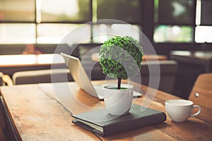 Small decorate tree, laptop, notebook and coffee cup in on wooden table in coffee shop. Relaxation and Business Co-working space