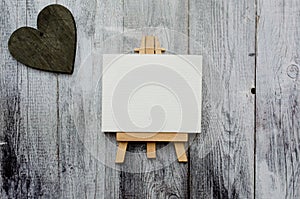 Small easel with a blank canvas over white and dark wooden heart. Old wooden background and big copy space for your sign