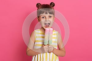 Small European kid biting fruit ice cream, little charming girl with widely opened mouth, wearing summer clothes, looks happy,