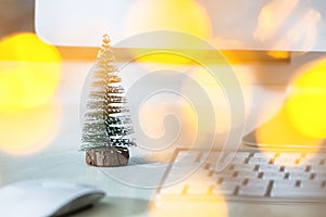 A small fir tree stands on a white desktop, near a computer monitor, keyboard and mouse. Concept, create a Christmas mood in