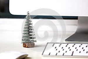 A small fir tree stands on a white desktop, near a computer monitor, keyboard and mouse. Concept, create a Christmas mood in
