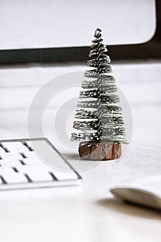 A small fir tree stands on a white desktop, near a computer monitor, keyboard and mouse. Concept, create a Christmas mood in the