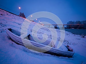 A small fishing boat on the shore covered with snow during a cold winter evening. Lights on the promenade by the river