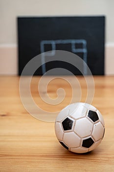 Small football on a wooden floor in focus. Abstract goal post drawn on a black board out of focus in the background. Soccer tactic