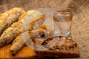 A small fried fish on a wooden board, pieces of bread and a glass of vodka, close-up, selective focus