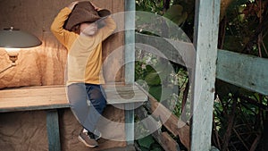 Small, funny child in big cowboy hat is sitting on wooden porch of country house