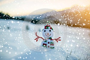 Small funny toy baby snowman in knitted hat and scarf in deep snow outdoor on blurred mountains landscape and falling big