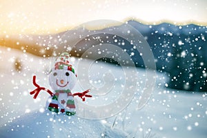 Small funny toy baby snowman in knitted hat and scarf in deep snow outdoor on blurred mountains landscape and falling big