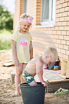 Small girl with blond curly hair and ittle boy play in the yard of the house in the sand.