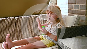 Small girl blowing soap bubbles on the balcony couch in summertime