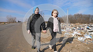 Small girl and boy holds hands of each other and goes along road to school against the background of garbage dump