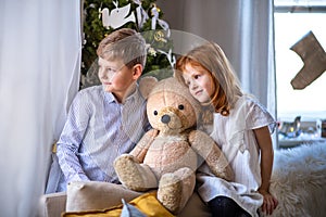 Small girl and boy indoors at home at Christmas, holding teddy bear.
