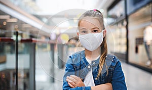 Small girl with face mask looking at camera indoors in shopping center, coronavirus concept.
