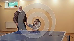 Small girl jumping near tennis table, holding rocket in hands. Grandfather comes and takes racket. Happy family play