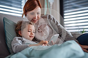 Small girl with mother in bed in hospital, using smartphone to pass time.
