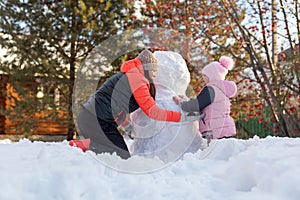 Small girl with mother sitting on snow making snowman with hands on backyard in evening with rowan and fir trees in