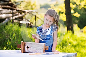 A small girl with a paintbrush outside, painting a wooden birdhouse.