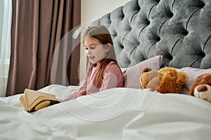 A small girl sitting in a bed wearing pyjamas covered with blanket reading her book and smiling with a teddy bear near