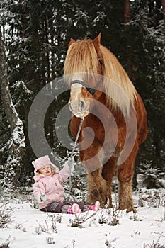 Small girl sitting in the snow and big palomino horse standing n