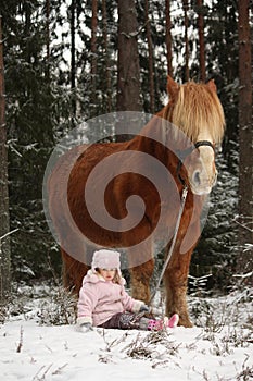 Small girl sitting in the snow and big palomino horse standing n