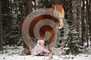 Small girl sitting in the snow and big palomino horse standing n