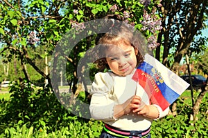 Small girl stands with flag of Russia in front of lilac Bush on sunny day