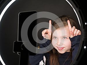 Small girl using camera of smartphone in front of ring light, shooting video for blog. Adorable child learning new technology.