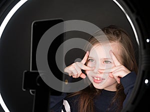 Small girl using camera of smartphone in front of ring light, shooting video for blog. Adorable child learning new technology.