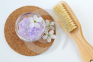 Small glass bowl with purple bath salt (foot soak), massage body brush and white flowers. Top view, copy space