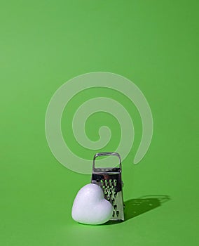 A small grater and a white heart on a green background. Harsh shadow. Text space. Brokenhearted, love, life, message, culinary photo