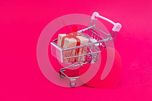 Small grocery cart with gift boxes on red-pink background. Give gifts with love on Valentine`s Day, Christmas and