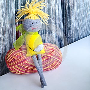 A small handmade doll sits with his legs crossed on a ball of colored woolen threads
