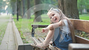 Small happy child girl sitting on a bench resting in summer park.