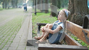 Small happy child girl sitting on a bench resting in summer park.