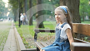 Small happy child girl sitting on a bench resting in summer park