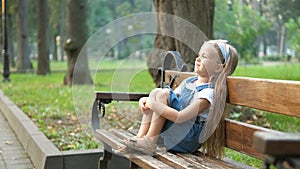 Small happy child girl sitting on a bench resting in summer park