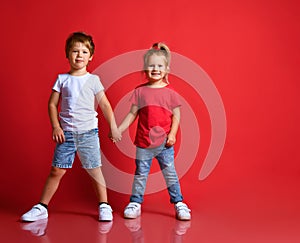 Small happy children boy and girl in stylish casual clothing standing, holding hands and feeling shy over red background