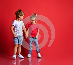 Small happy children boy and girl in stylish casual clothing standing, holding hands and feeling shy over red background