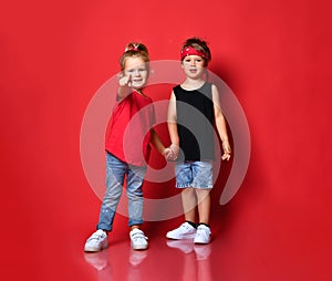 Small happy children boy and girl in stylish casual clothing standing, holding hands and pointing at camera over red background