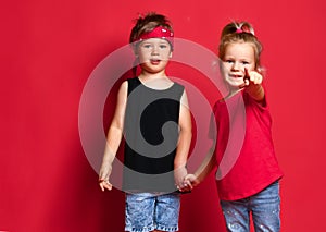 Small happy children boy and girl in stylish casual clothing standing, holding hands and pointing at camera over red background