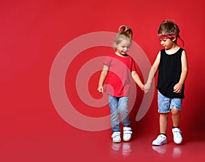 Small happy cute children boy and girl in stylish casual clothing go holding hands as couple over red background