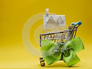 Small house with glitter in a metal shopping cart with green bow and Irish shamrock on a side, yellow background. Buying home in