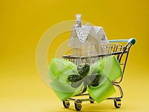 Small house with glitter in a metal shopping cart with green bow and Irish shamrock on a side, yellow background. Buying home in
