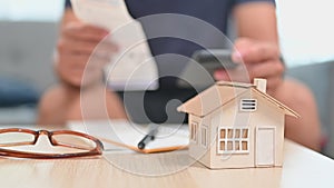 Small house model on table with man using smartphone for making online payments, managing expenses finances in living