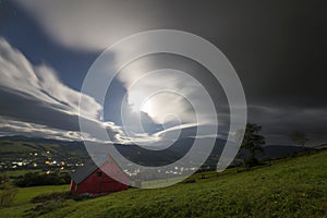 Small house on mountain slope under cloudy sky, bright lights in photo