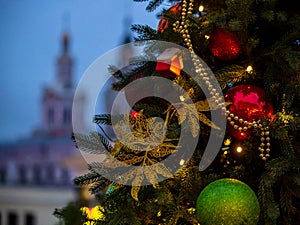 A small house, red and green balls, yellow beads and golden snowflakes hang on a Christmas tree outdoors